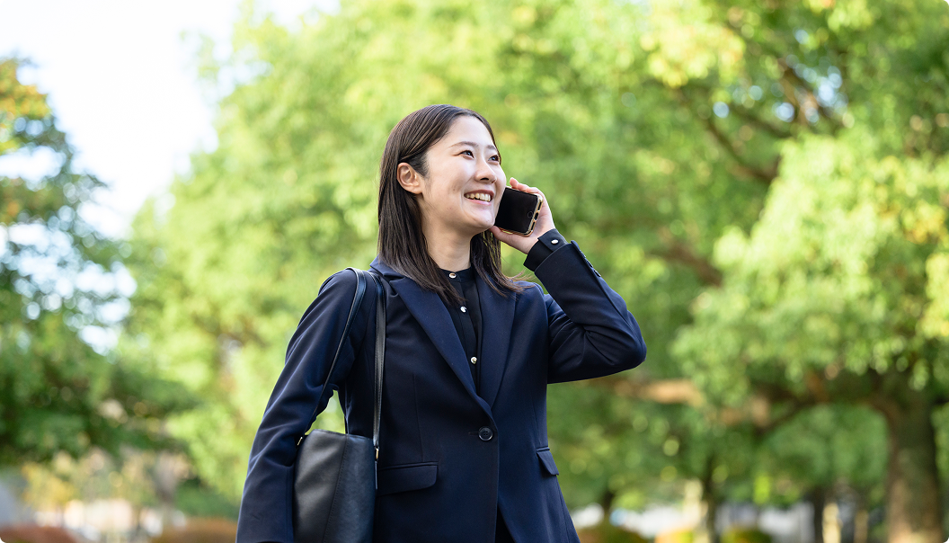 スーツを着た女性が緑の多い屋外で電話をしながら笑顔で歩いている