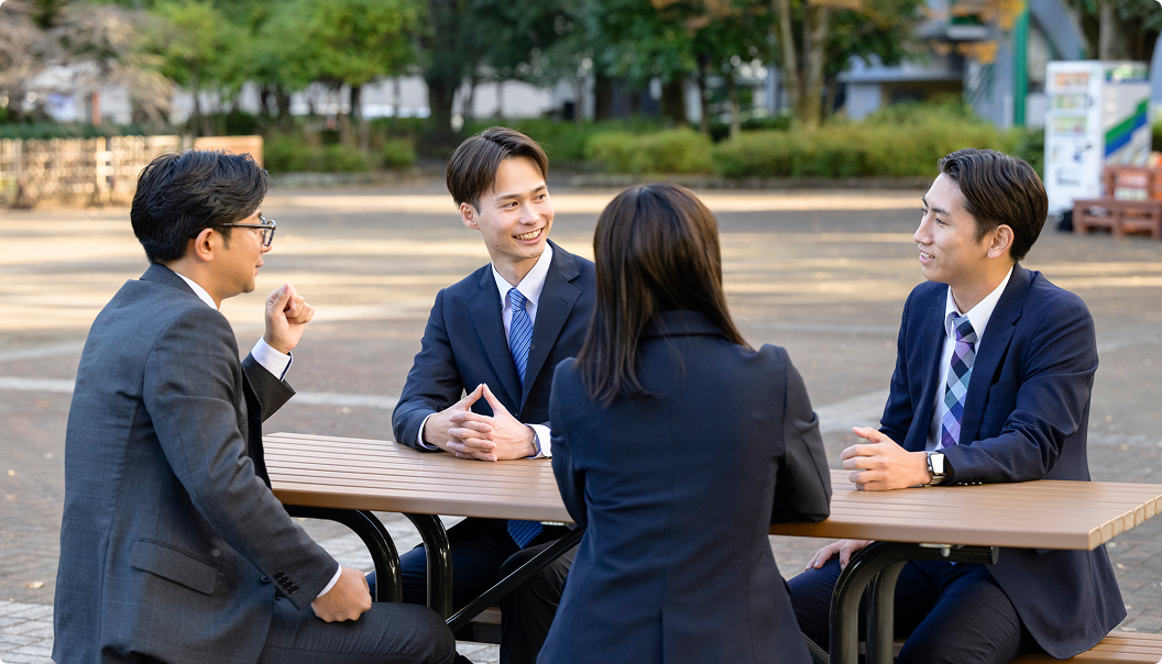 スーツ姿の若手ビジネスパーソン4人が屋外のベンチで談笑している様子