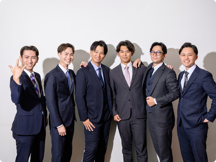 スーツ姿の男性6人が横一列に並び、肩を組み笑顔でポーズを取る集合写真。白い壁を背景にしたスタジオ風の撮影