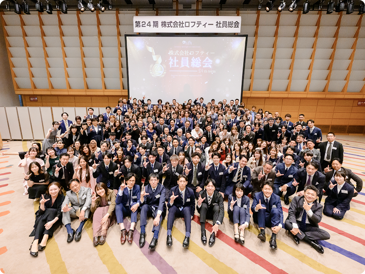 大規模ホールで大勢の参加者が前列に座り、全員でポーズを取る社員総会の集合写真