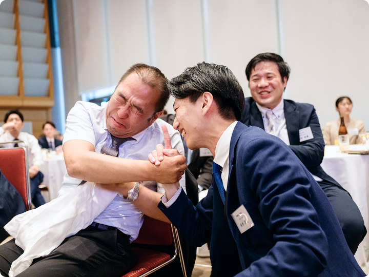 椅子に座った男性2人が腕相撲をし、周囲の参加者が笑顔で見守る余興の場面