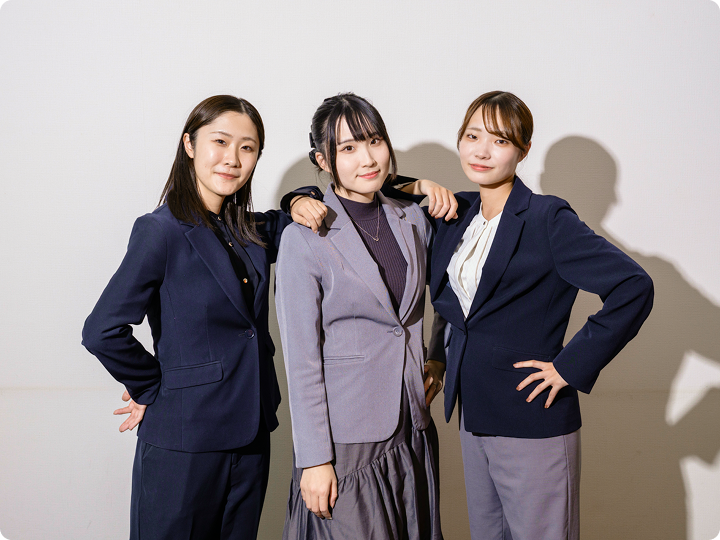 スーツ姿の女性3人が並び、腕を組んで自信のある表情を見せるスタジオ風写真