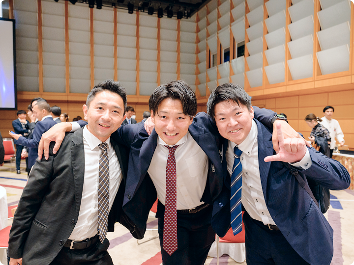 スーツ姿の男性3人が肩を組み、前かがみで笑顔を見せる懇親会中の写真