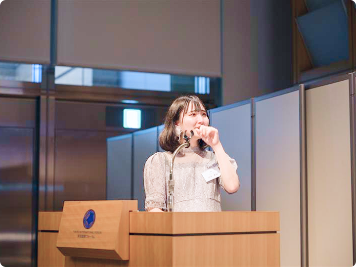 演台の前に立つ女性がマイクでスピーチをし、少し照れた表情を見せる場面